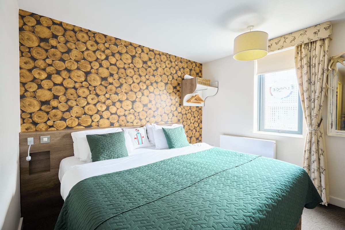A hotel room with a bed, a wooden log wall, a window with curtains, and a bedside lamp.