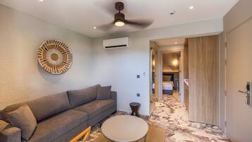 Una habitación de hotel moderna con un sofá gris, una mesa redonda, un ventilador de techo y un pasillo que conduce a un baño.