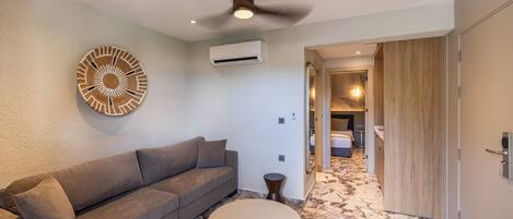 Ein modernes Hotelzimmer mit grauem Sofa, rundem Tisch, Deckenventilator und Flur zum Badezimmer.