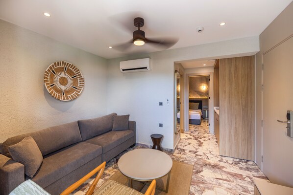 Quarto de hotel moderno com sofá cinzento, mesa redonda, ventoinha no teto e corredor que dá para um banheiro.
