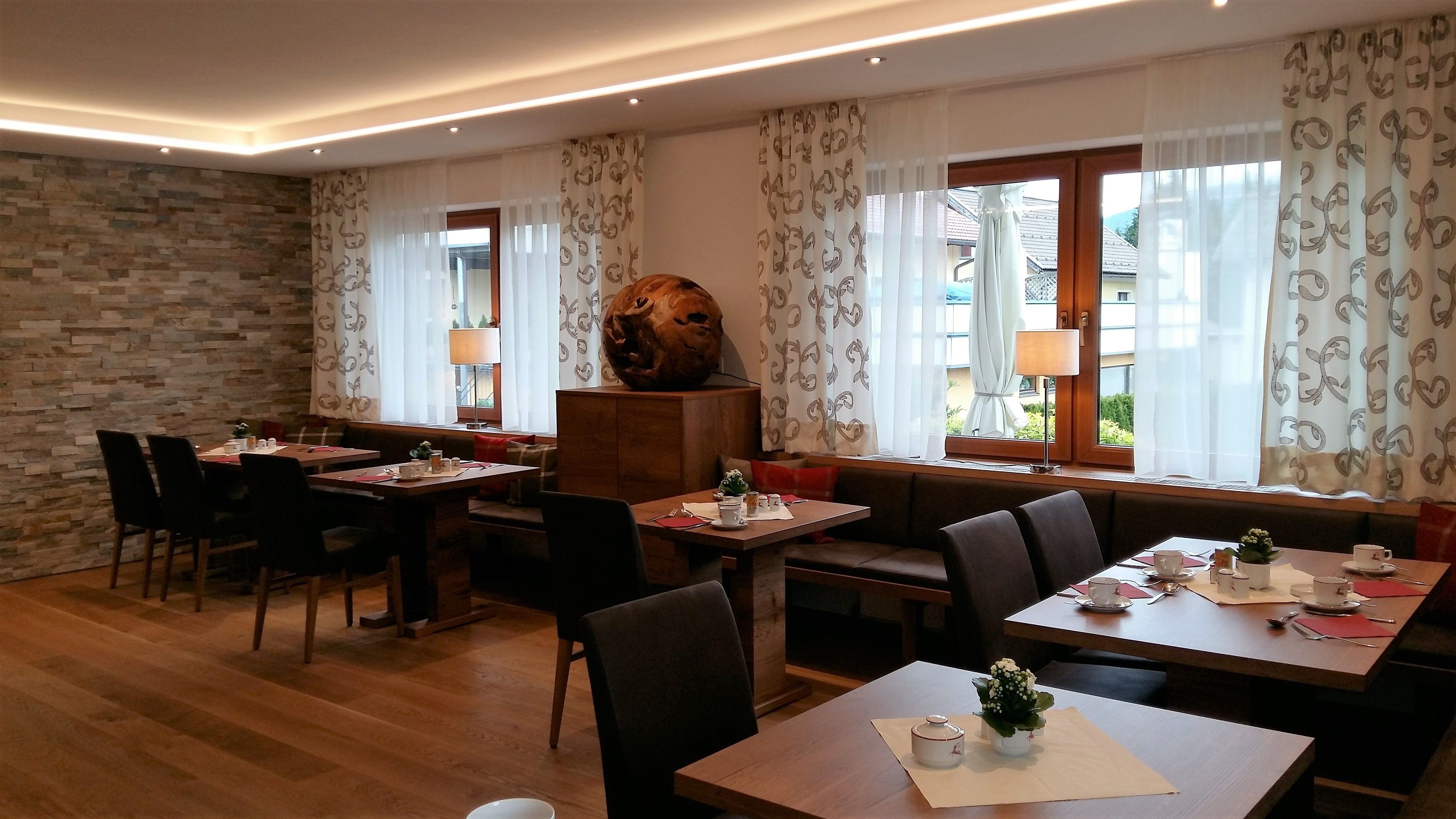 A dining area with wooden tables and chairs, a stone wall, and large windows with patterned curtains.