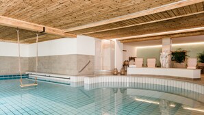 A large indoor swimming pool with a wooden ceiling, a stone wall, and a reception area with a statue and plants. - Landhaus Steiner (Altenmarkt im Pongau)
