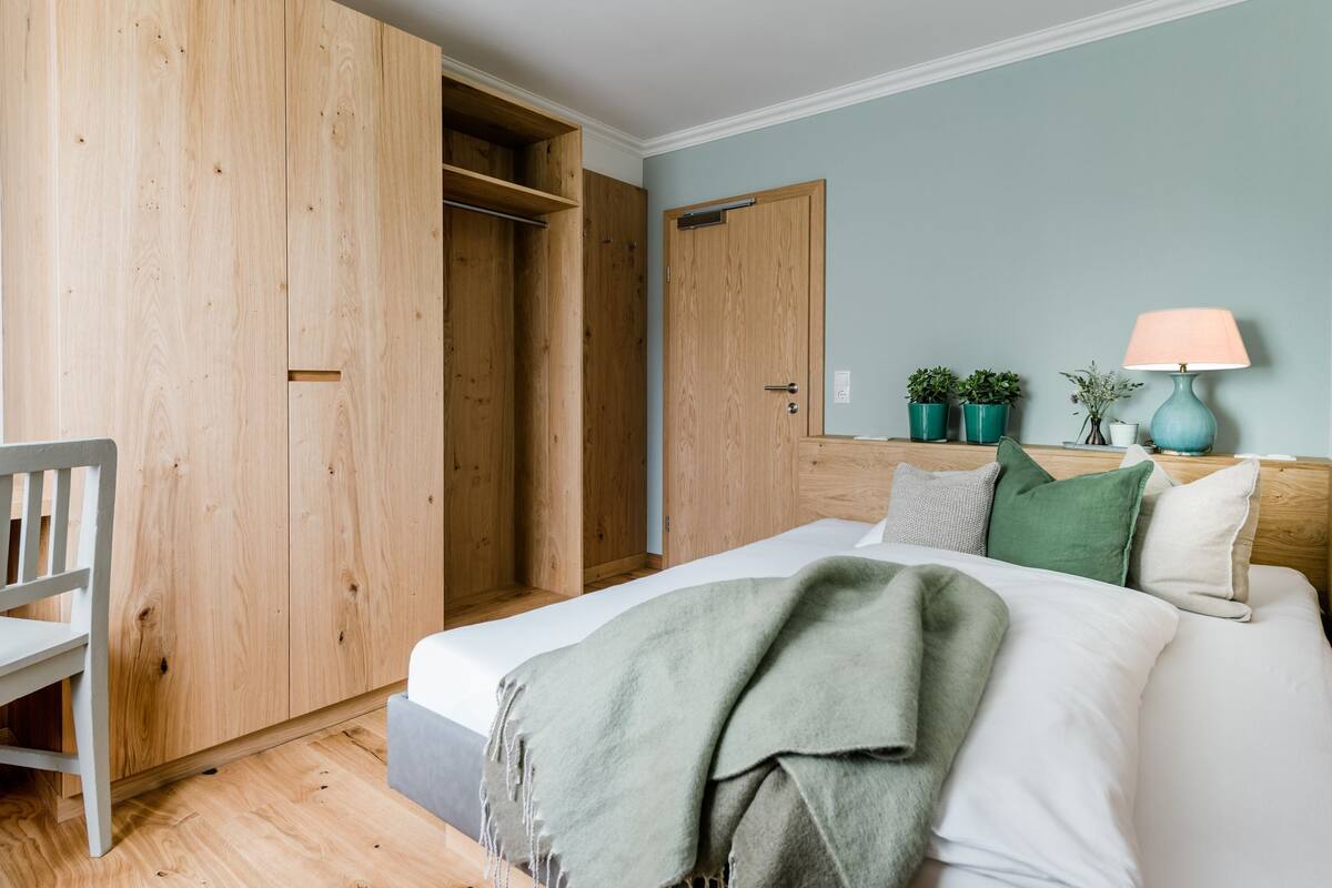 A bedroom with a bed, a wooden wardrobe, a chair, and a bedside table with a lamp.