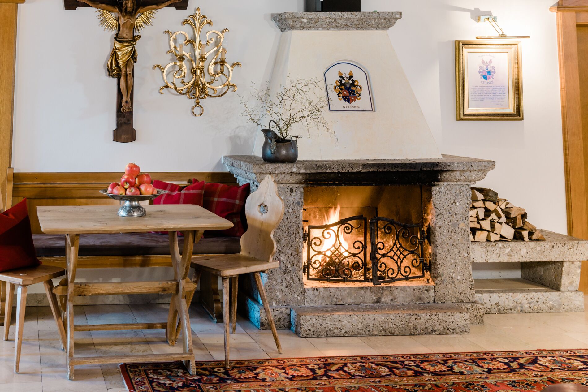 A cozy living room with a stone fireplace, a wooden bench, a table with a fruit bowl, and a chandelier.