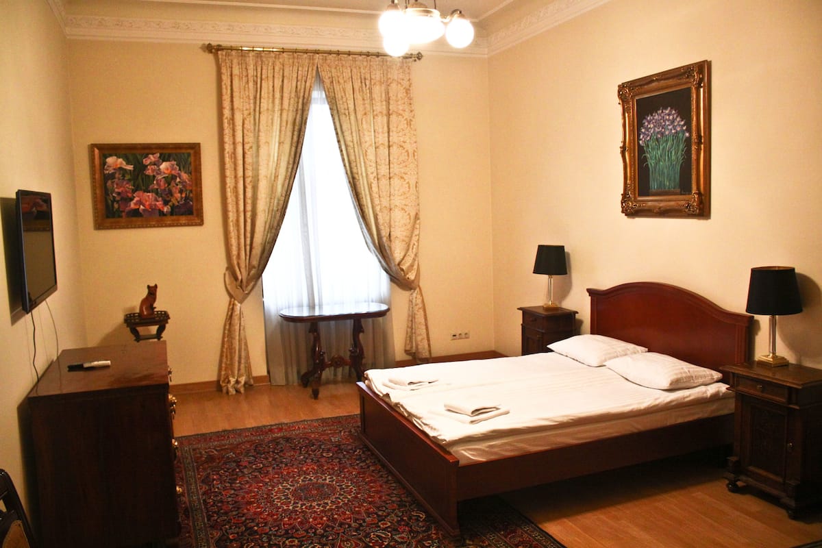 Un dormitorio con una cama de madera, una mesita de noche, un escritorio, una silla, una ventana con cortinas y dos cuadros en la pared.