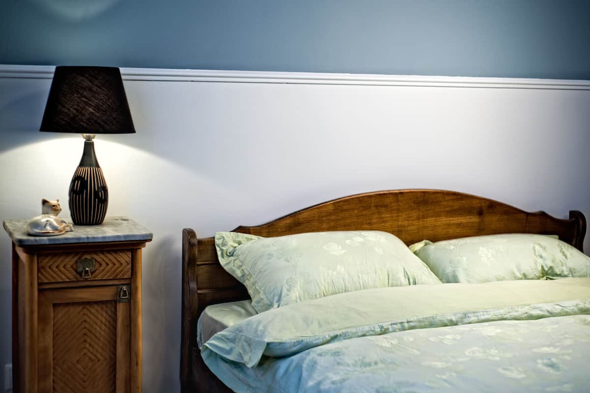 Un dormitorio con cabecera de madera, mesita de noche con lámpara y una figurita decorativa de un gato.
