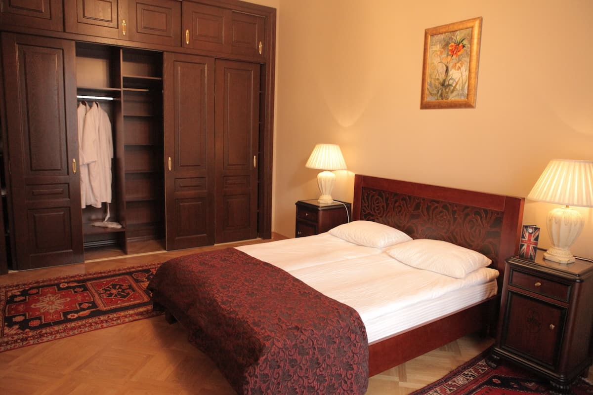 Un dormitorio con un amplio armario de madera, una cama con sábanas blancas, dos mesitas de noche con lámparas y una alfombra decorativa.