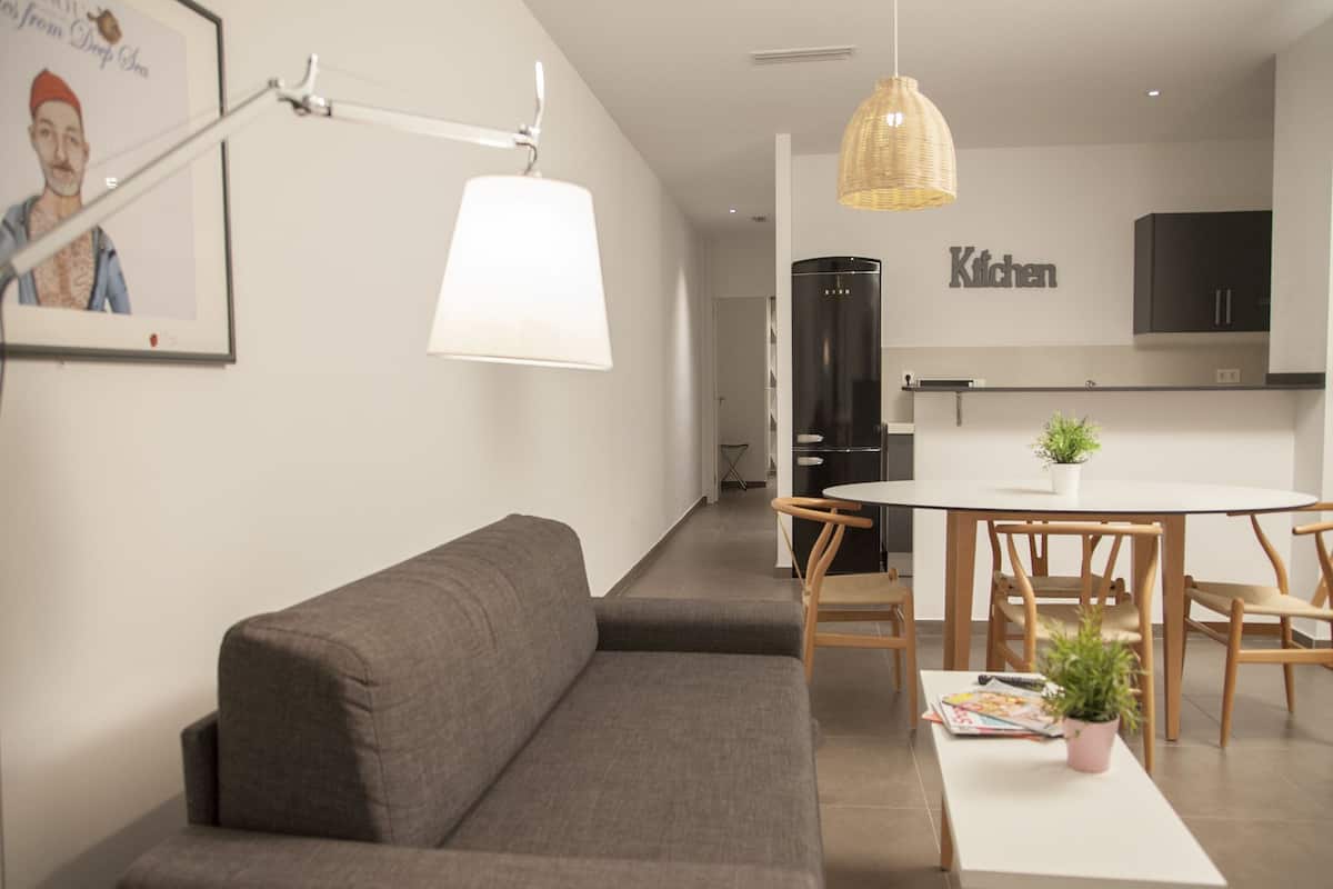 A modern living room with a grey sofa, a white table with a magazine and a plant, and a kitchen area in the background with a black refrigerator and a 'Kitchen' sign.