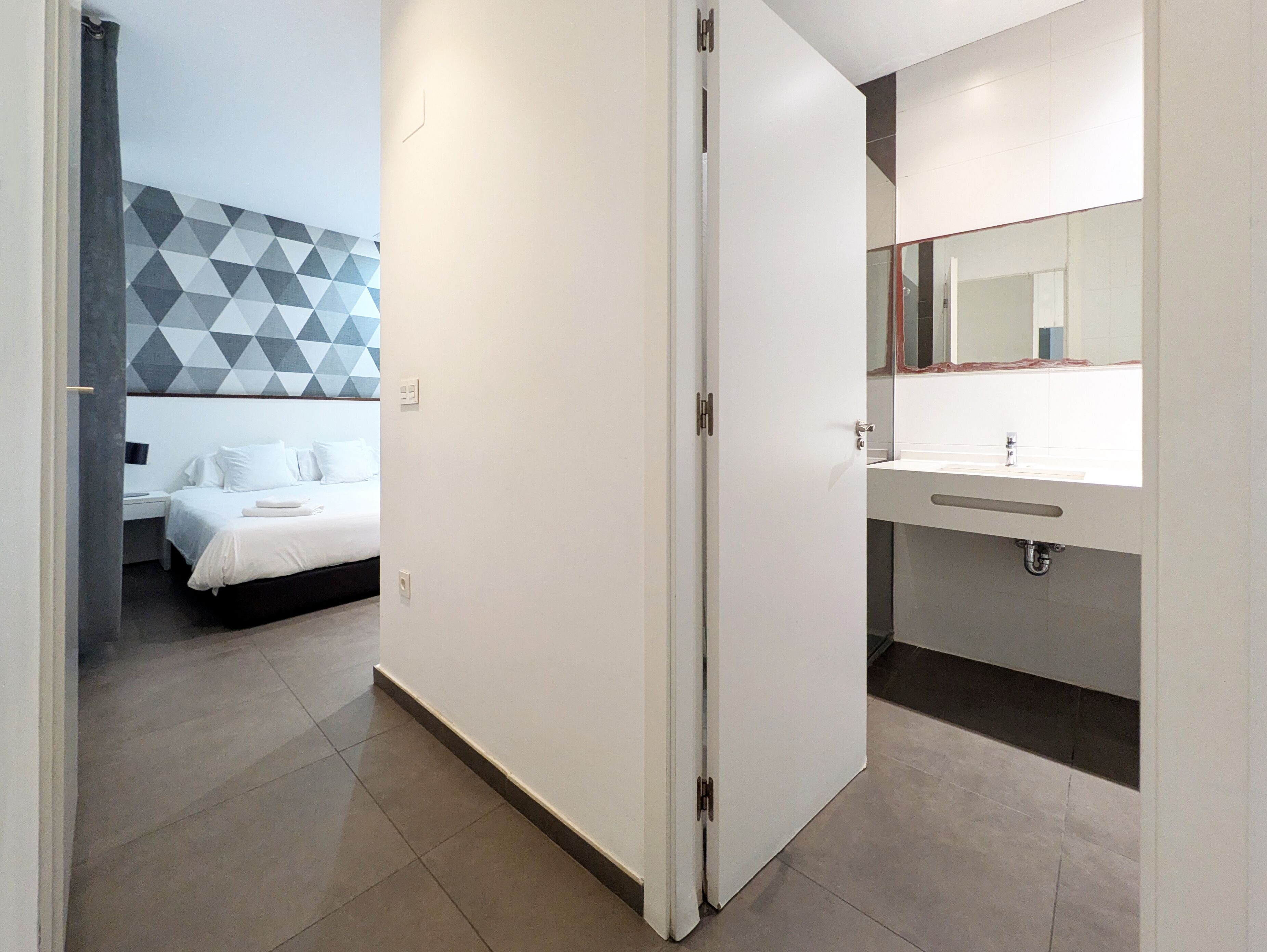 A hotel room with a bed, a bathroom with a mirror and sink, and a geometric-patterned wall.