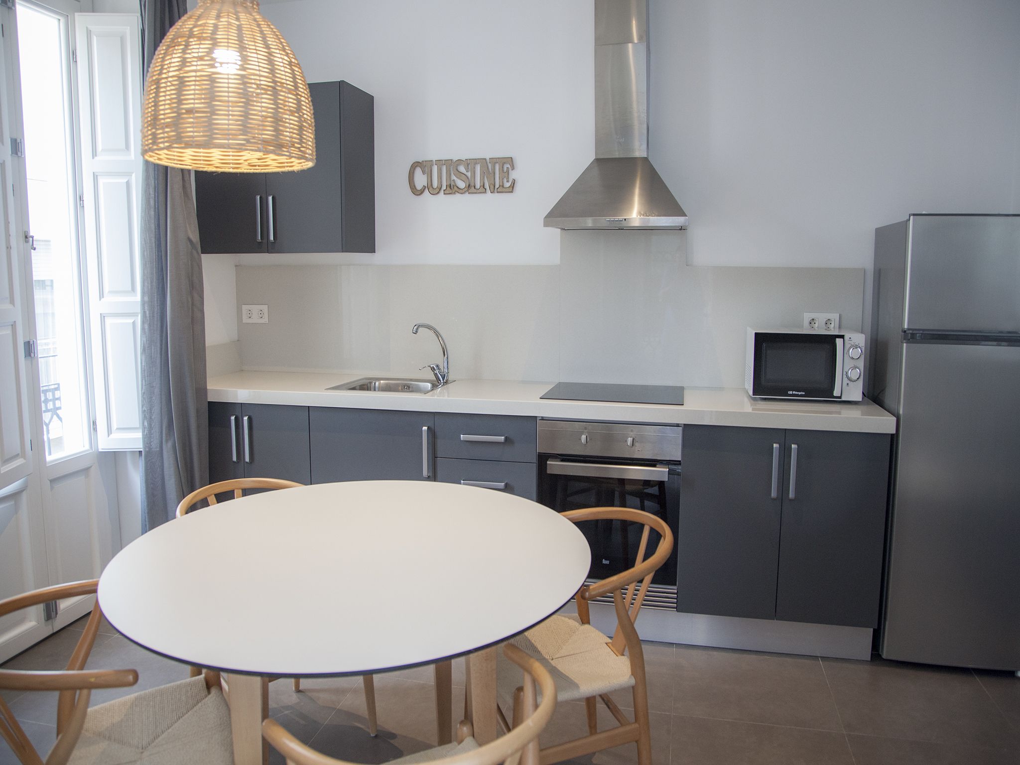 Une cuisine moderne avec des meubles gris, une hotte en acier inoxydable, un four à micro-ondes et un four encastré. On y trouve une table ronde avec des chaises et un luminaire suspendu en osier.