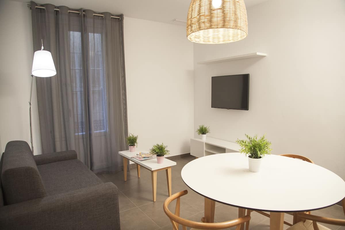 A modern living room with a grey sofa, a round white table with chairs, a small white side table with two potted plants, a flat-screen TV mounted on the wall, and a large window with grey curtains.