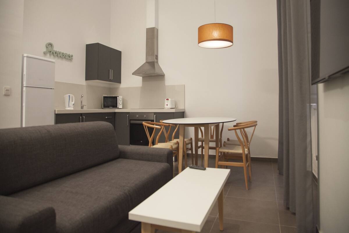A modern living room with a grey sofa, a white table, and a kitchen area with a microwave, kettle, and cabinets.