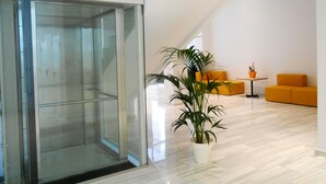 Un interior moderno con un ascensor de vidrio, una planta en maceta y asientos color naranja.
