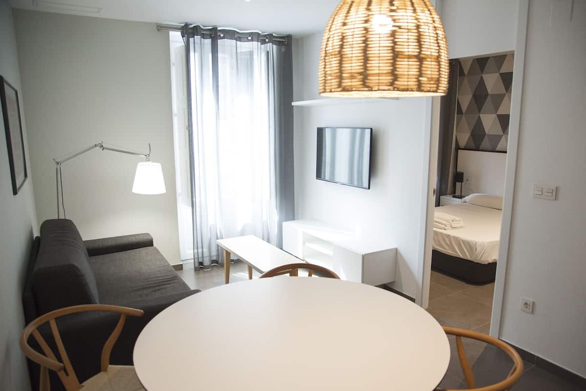 A compact hotel room with a round dining table, two chairs, a sofa, a TV mounted on the wall, and a bed with a geometric-patterned headboard.