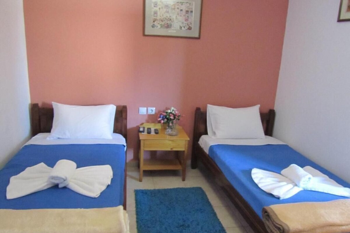 Une chambre avec deux lits simples, chacun recouvert d’une couvre-lit bleue et agrémenté d’oreillers blancs, une petite table de chevet en bois sur laquelle est posé un vase de fleurs, et un tapis bleu sur le sol.