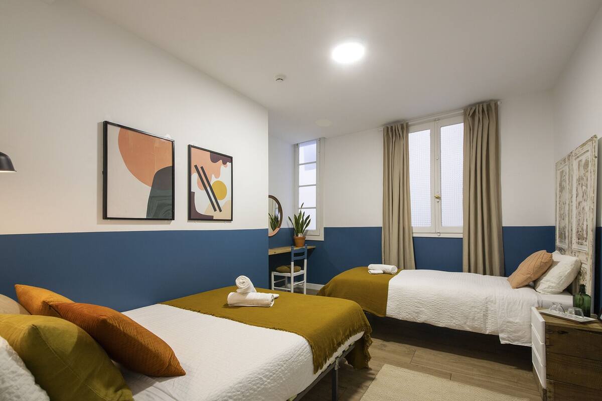 Quarto de hotel com duas camas, uma escrivaninha, uma cadeira, uma planta em vaso e obras de arte na parede.