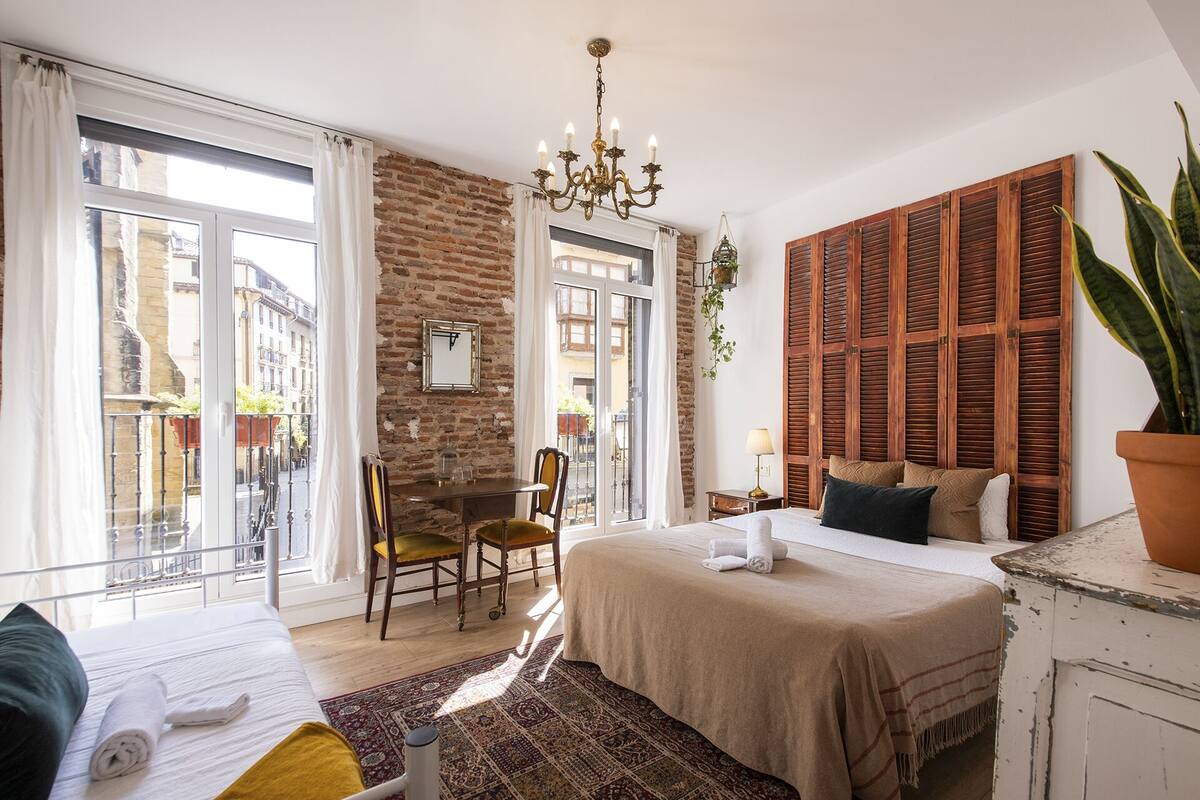 Quarto com cama, escrivaninha, cadeira, lustre e vista para a rua.