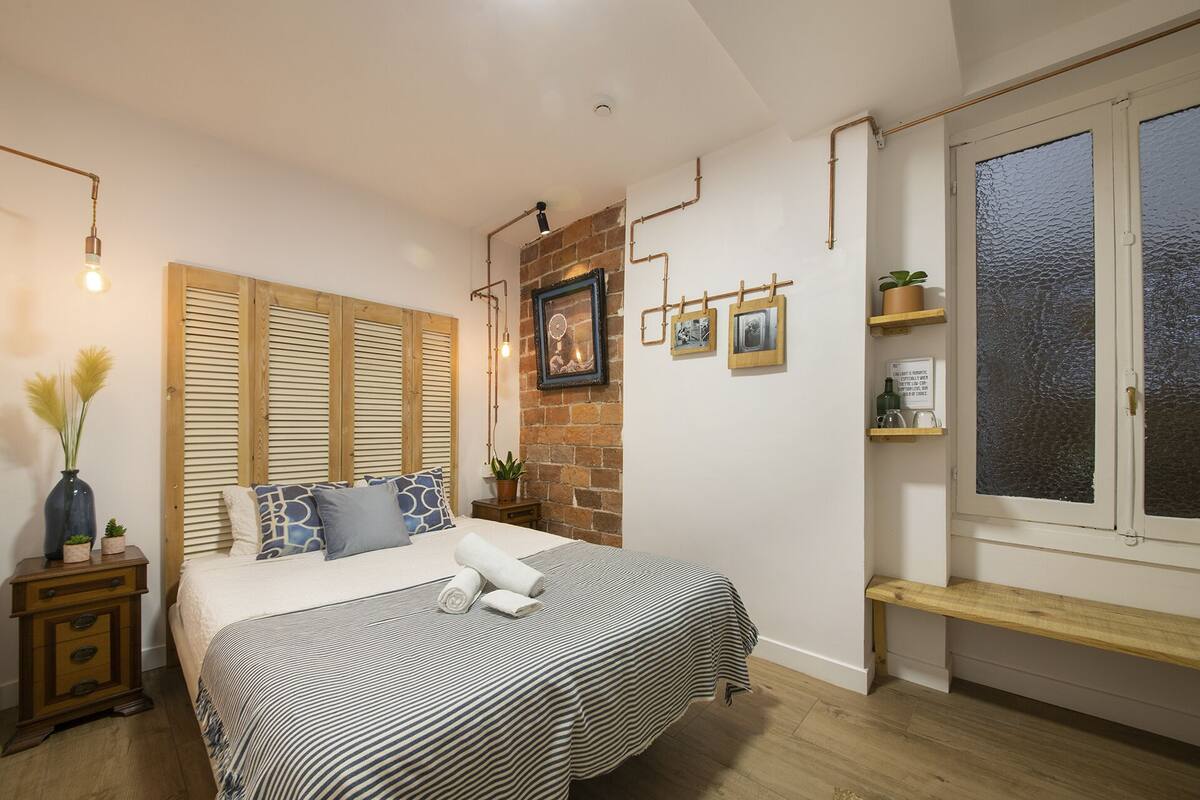 Quarto com cama, mesa de cabeceira, janela e parede de tijolos.