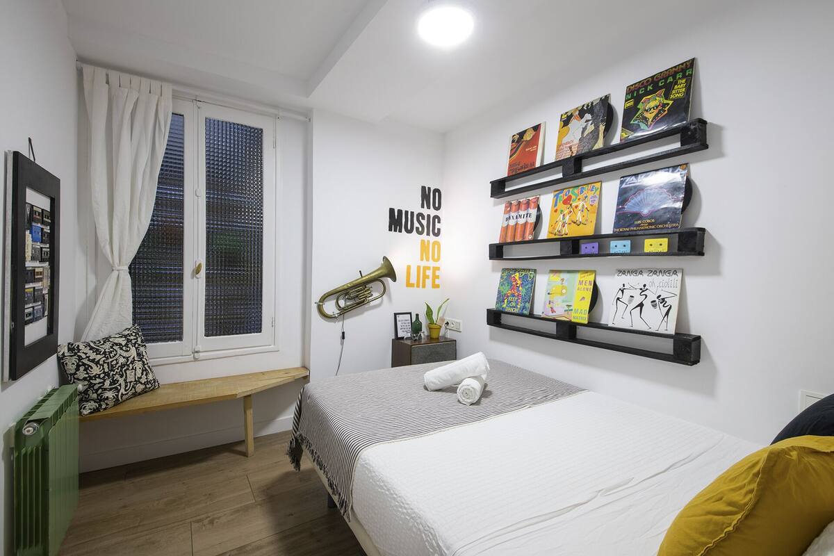 Quarto moderno com cama, janela com cortinas, prateleira com livros e um aviso na parede que diz 'NO MUSIC NO LIFE'.