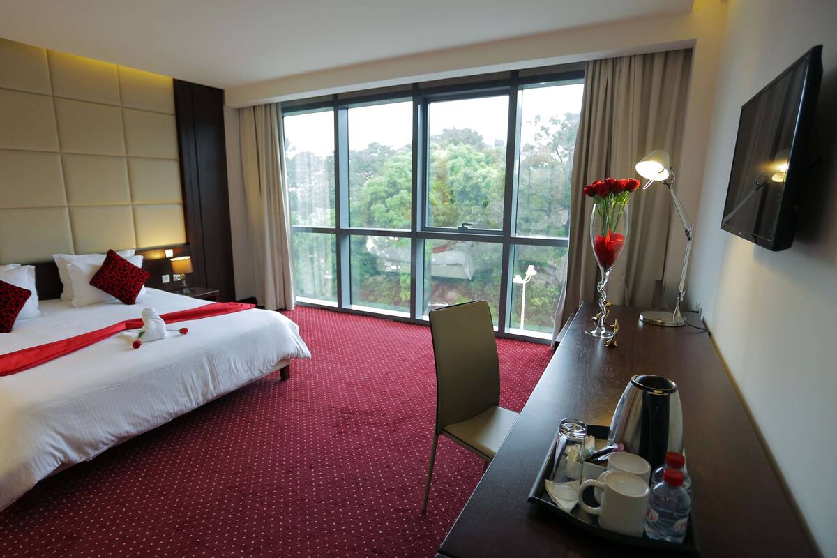 A hotel room with a large bed, a desk with a coffee maker, and a view of greenery through the window.