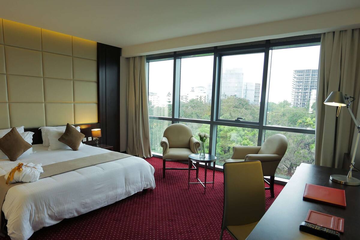 A hotel room with a large bed, two armchairs, a desk with a lamp, and a view of the city through large windows.