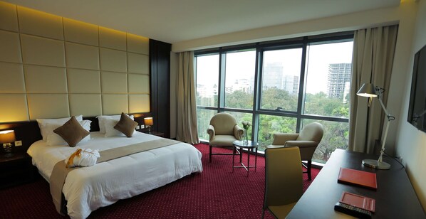 A hotel room with a large bed, two armchairs, a desk with a lamp, and a view of the city through large windows.