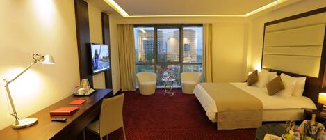 A hotel room with a bed, a desk with a TV, a chair, and a view of buildings through large windows.