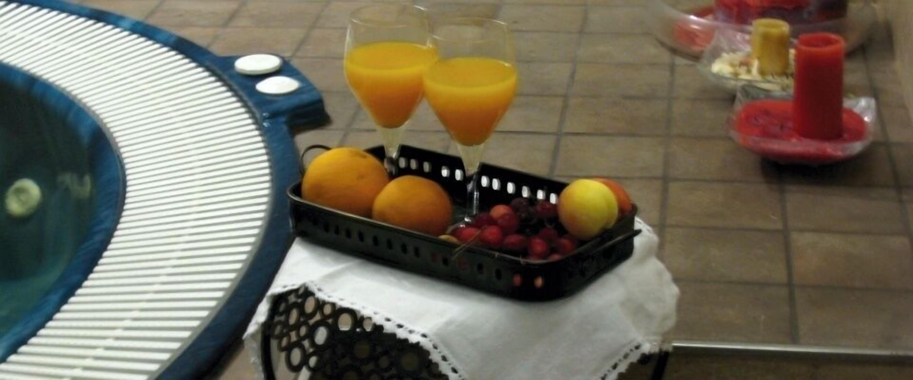 Disposição para o café da manhã com uma cesta de frutas, dois copos de suco de laranja e uma banheira de hidromassagem.