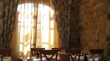 Un comedor con paredes de piedra, una mesa dispuesta para cuatro personas con copas de vino y cortinas con motivos florales.