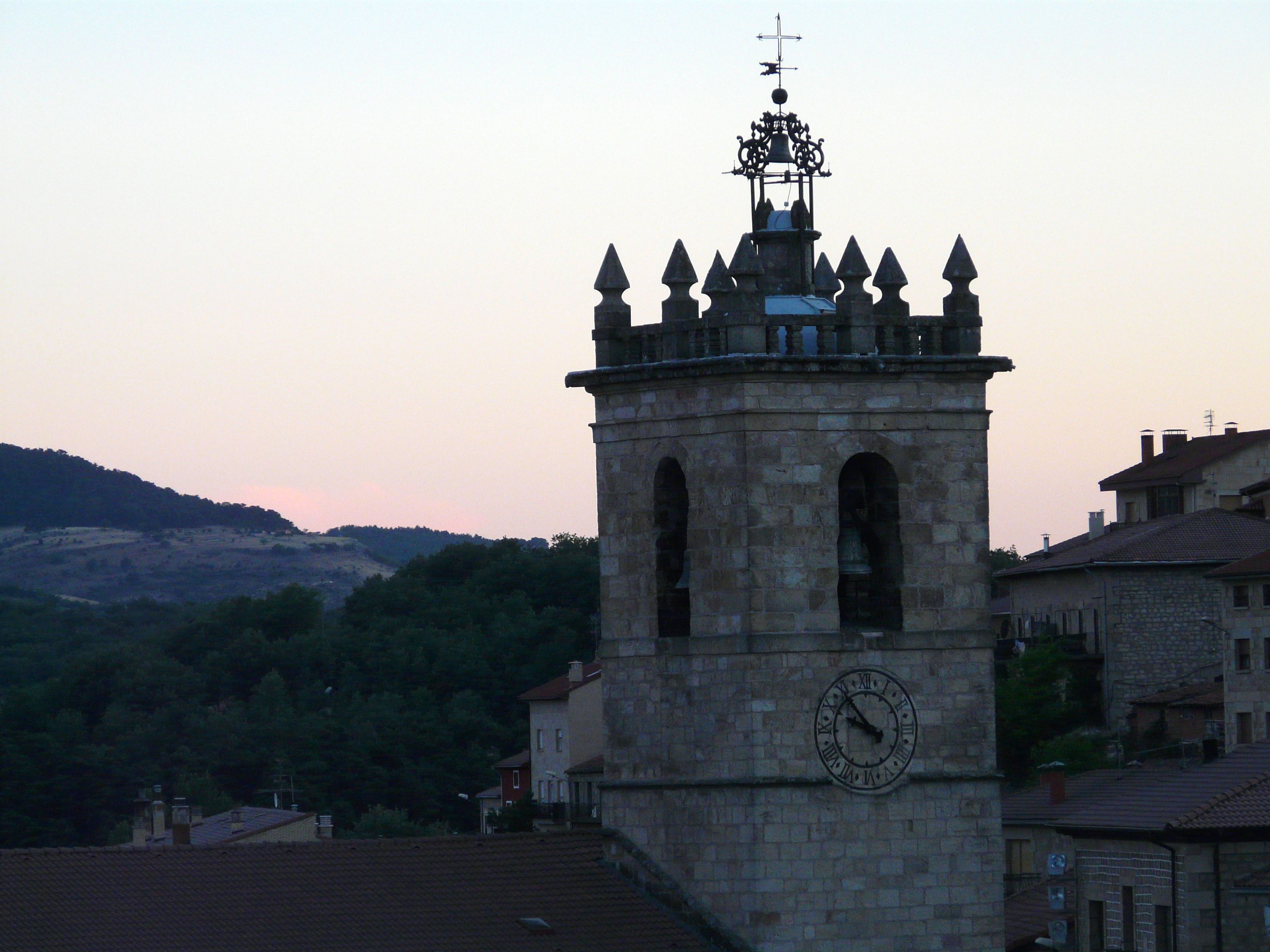 A stone clock tower with a weather vane on top, set against a backdrop of hills and residential buildings.