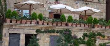 Edifício com mesas ao ar livre sob guarda-sóis, plantas em vasos na varanda e fachada de pedra.