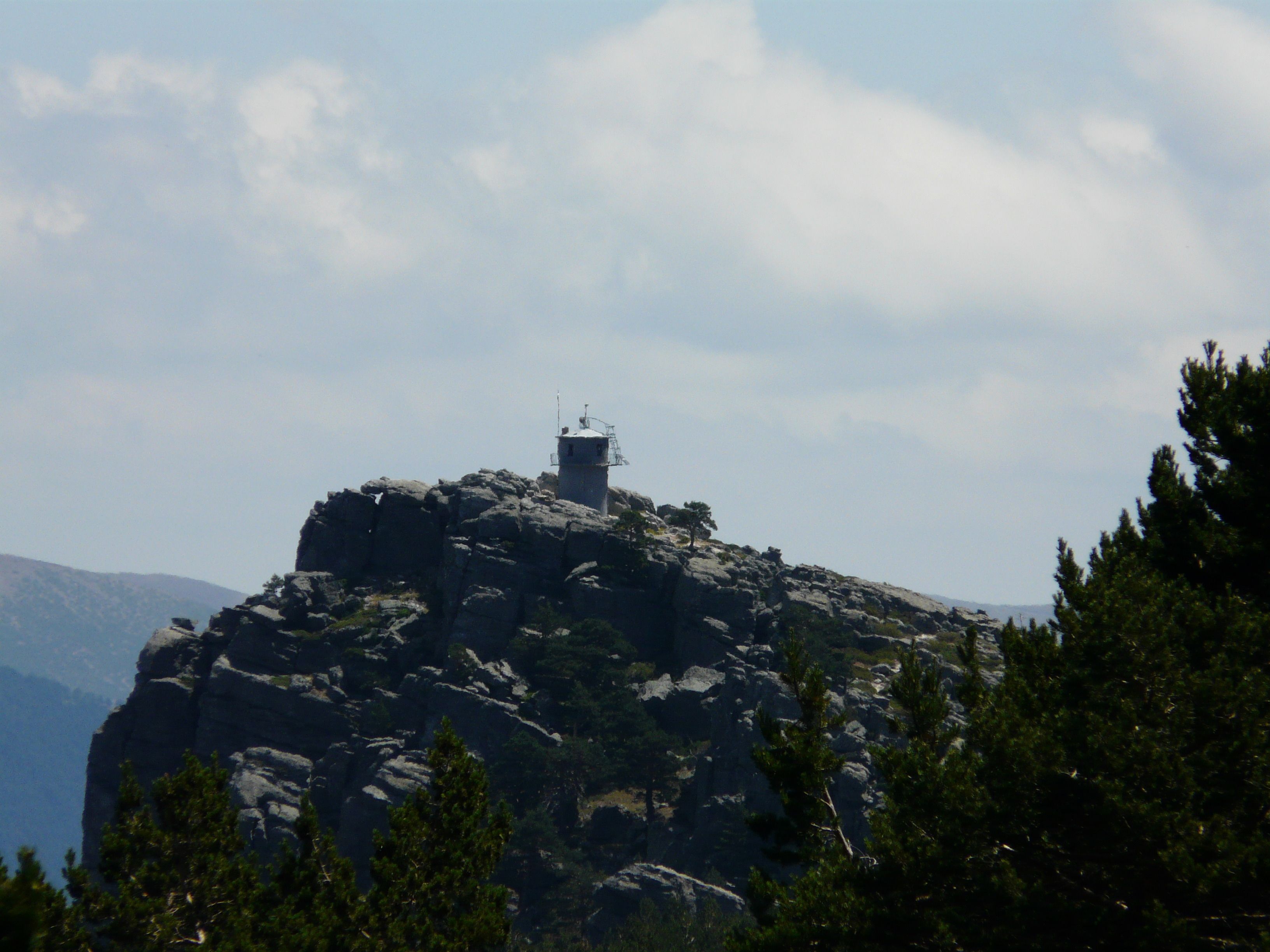 Un pico montañoso rocoso con una pequeña torre en la cima, rodeado de árboles.