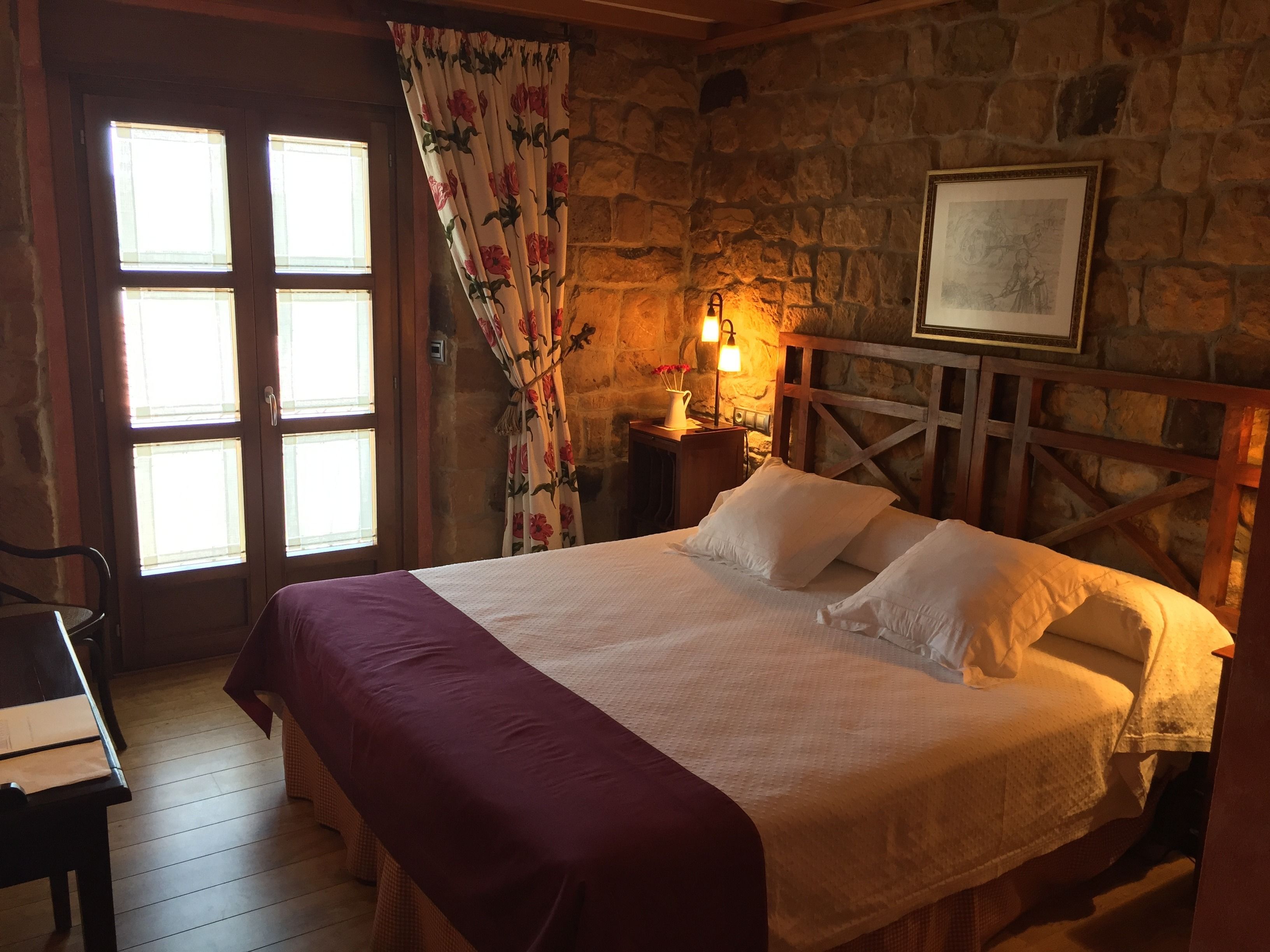 Una habitación con cama, cabecera de madera, pared de piedra, ventana con cortinas y mesita de noche con lámpara.