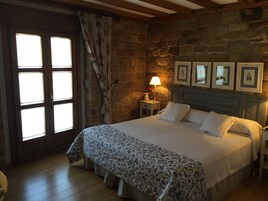 Quarto com parede de pedra, cama com colcha estampada, mesa de cabeceira com abajur e porta com cortina florida.