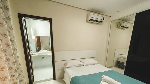 A hotel room with a bed, a bathroom visible through an open door, and an air conditioning unit on the wall. - MDJ Suites (Cabo Frio)