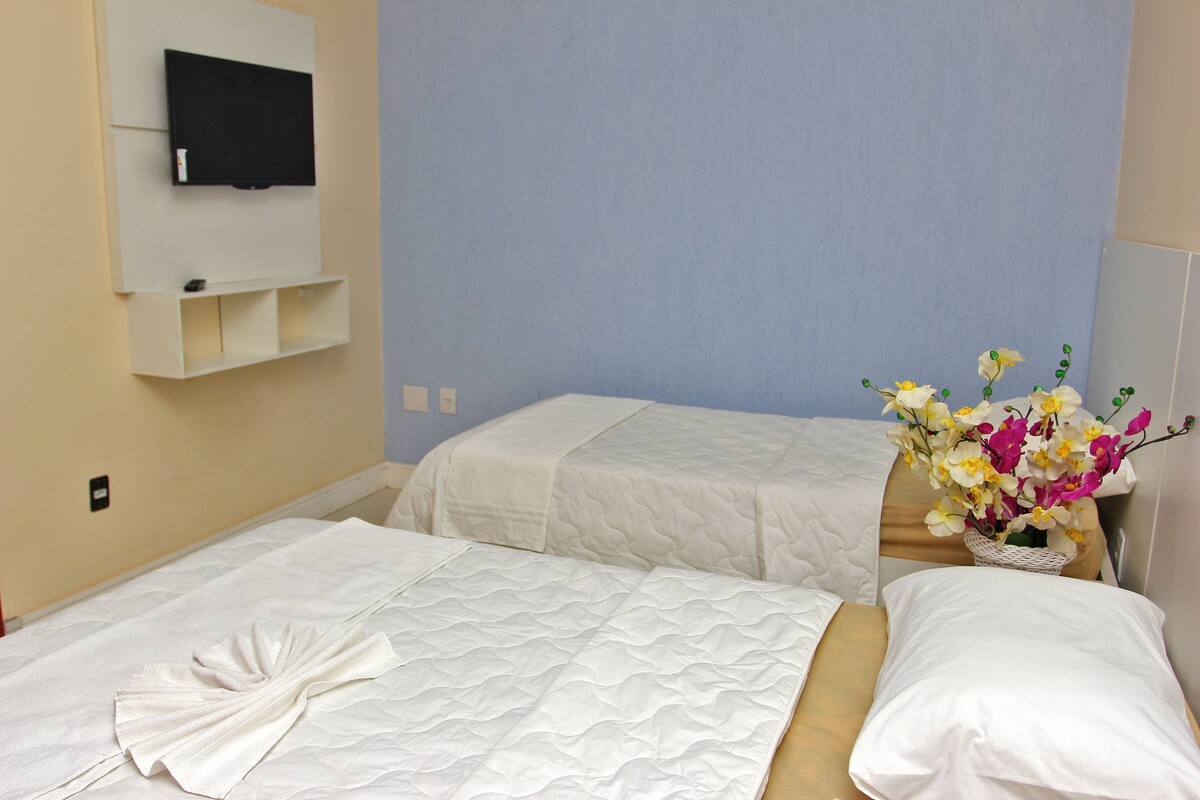 Quarto de hotel com duas camas, uma televisão de tela plana na parede e uma pequena mesa com um vaso de flores.