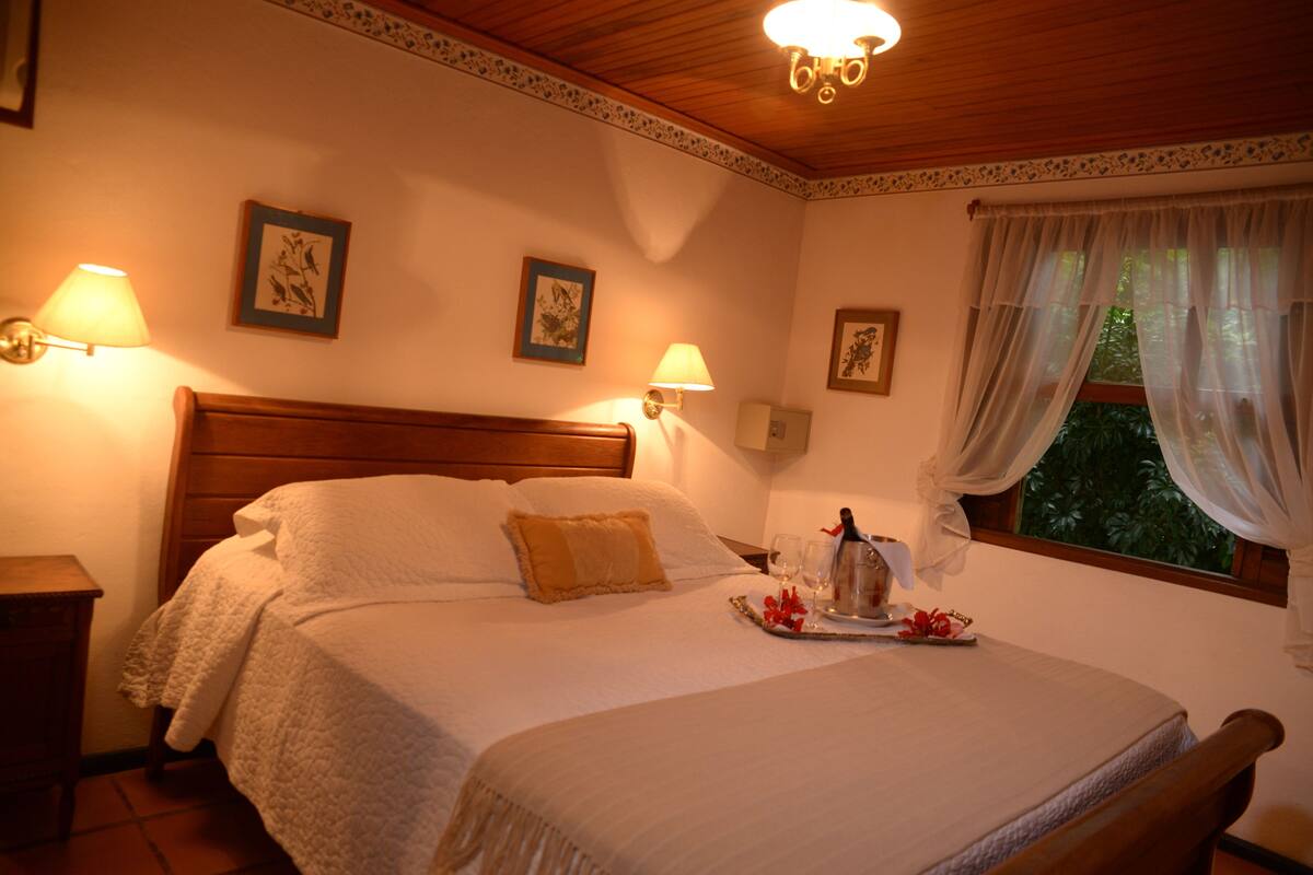 Un dormitorio bien iluminado con cabecera de madera, cama con ropa de cama blanca, dos lámparas fijadas a la pared y una ventana con cortinas translúcidas.