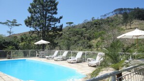 Piscina con sillas de descanso y sombrillas, rodeada por una valla y árboles.