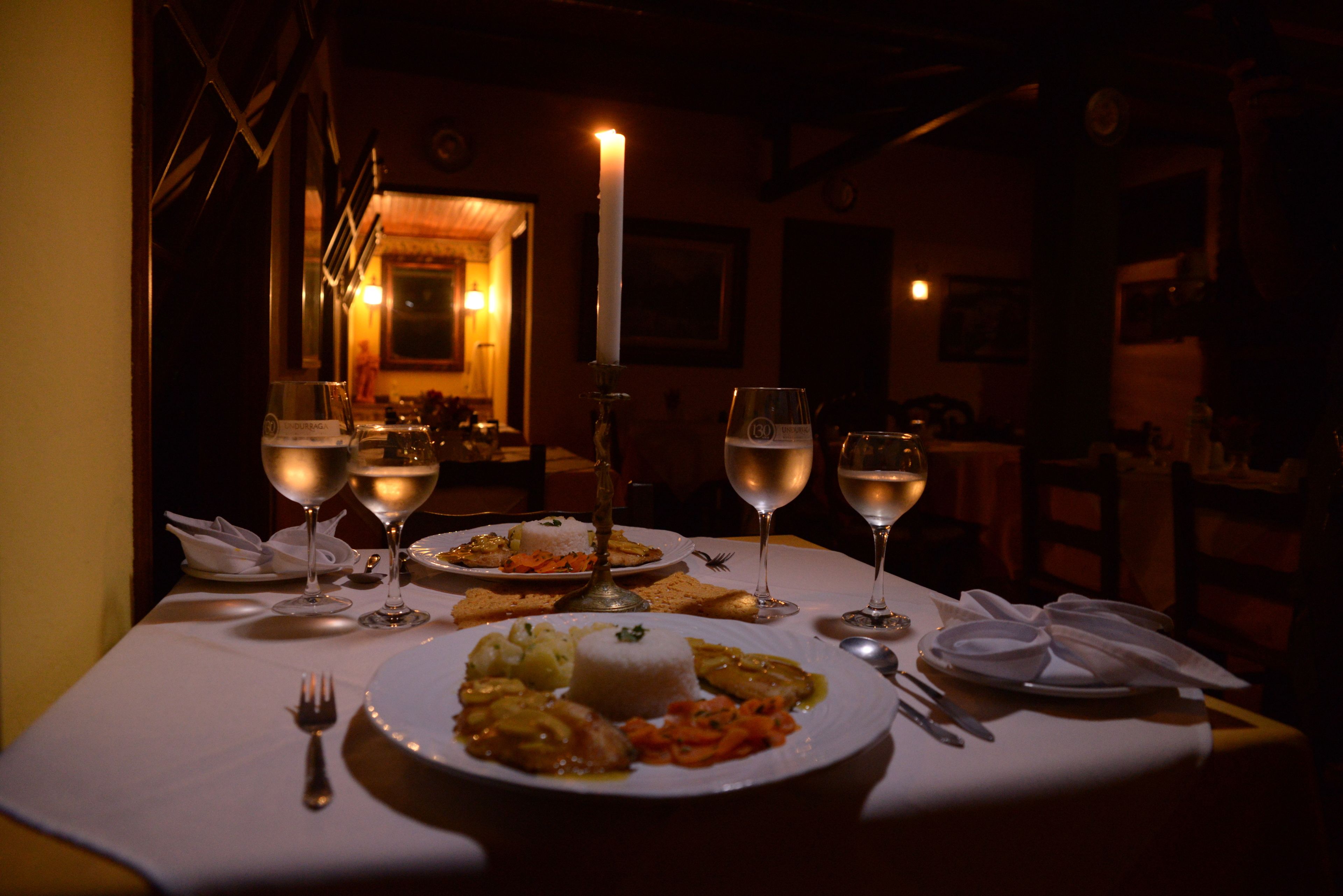 Una mesa de comedor tenuemente iluminada, con mantel blanco, platos de comida y copas de vino.
