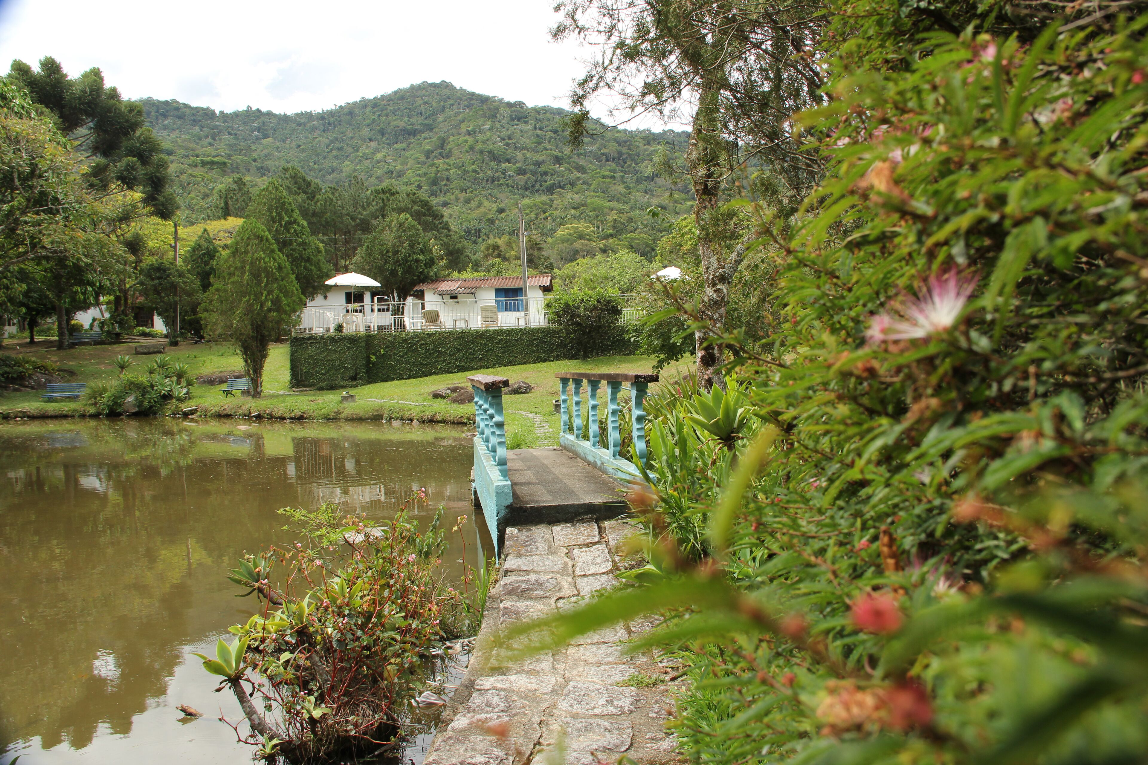 A serene lakeside setting with a stone bridge leading to a white building with a blue roof, surrounded by lush greenery and hills in the background.