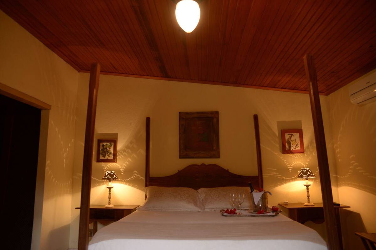 Una cama con dosel y tablero de madera, dos mesitas de noche con lámparas y una bandeja con una copa de vino y fresas sobre la cama.