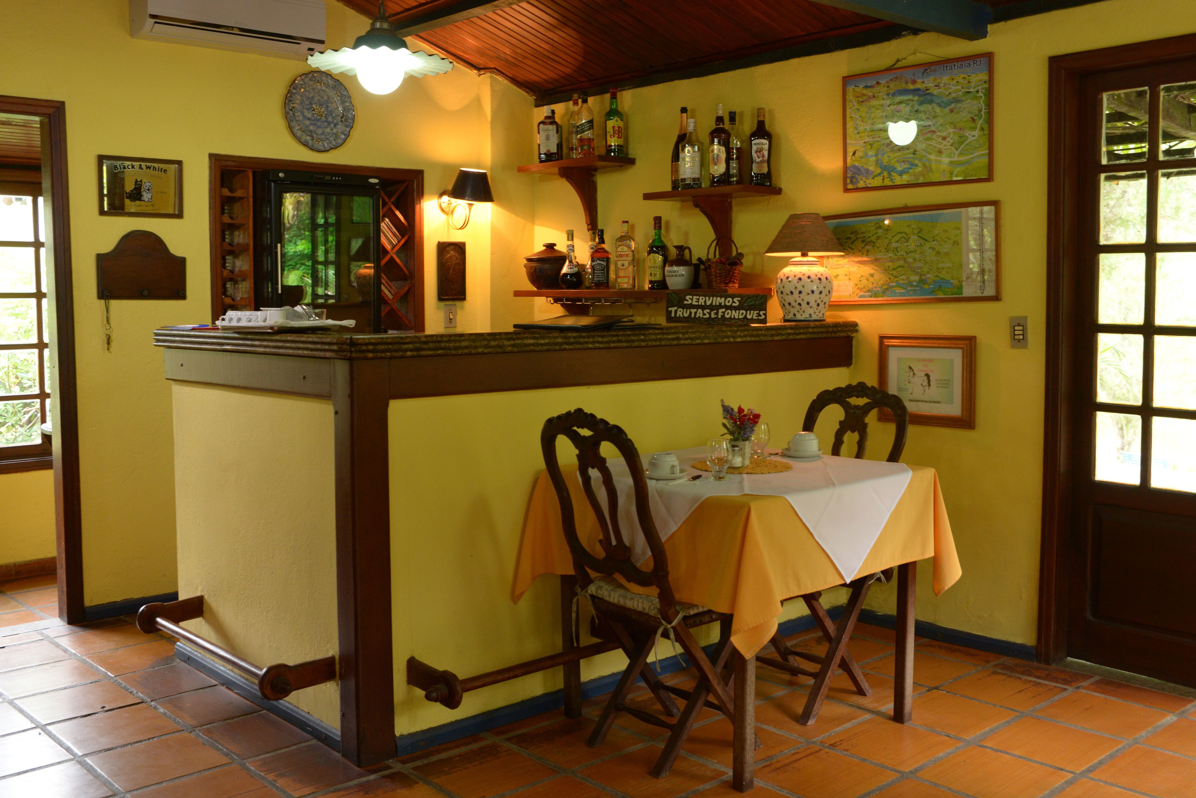 A bar area with a counter, chairs, and a table with a tablecloth. There are bottles and a lamp on the counter.