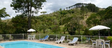 A swimming pool with lounge chairs and umbrellas, surrounded by a fence and trees, with a hilly landscape in the background.