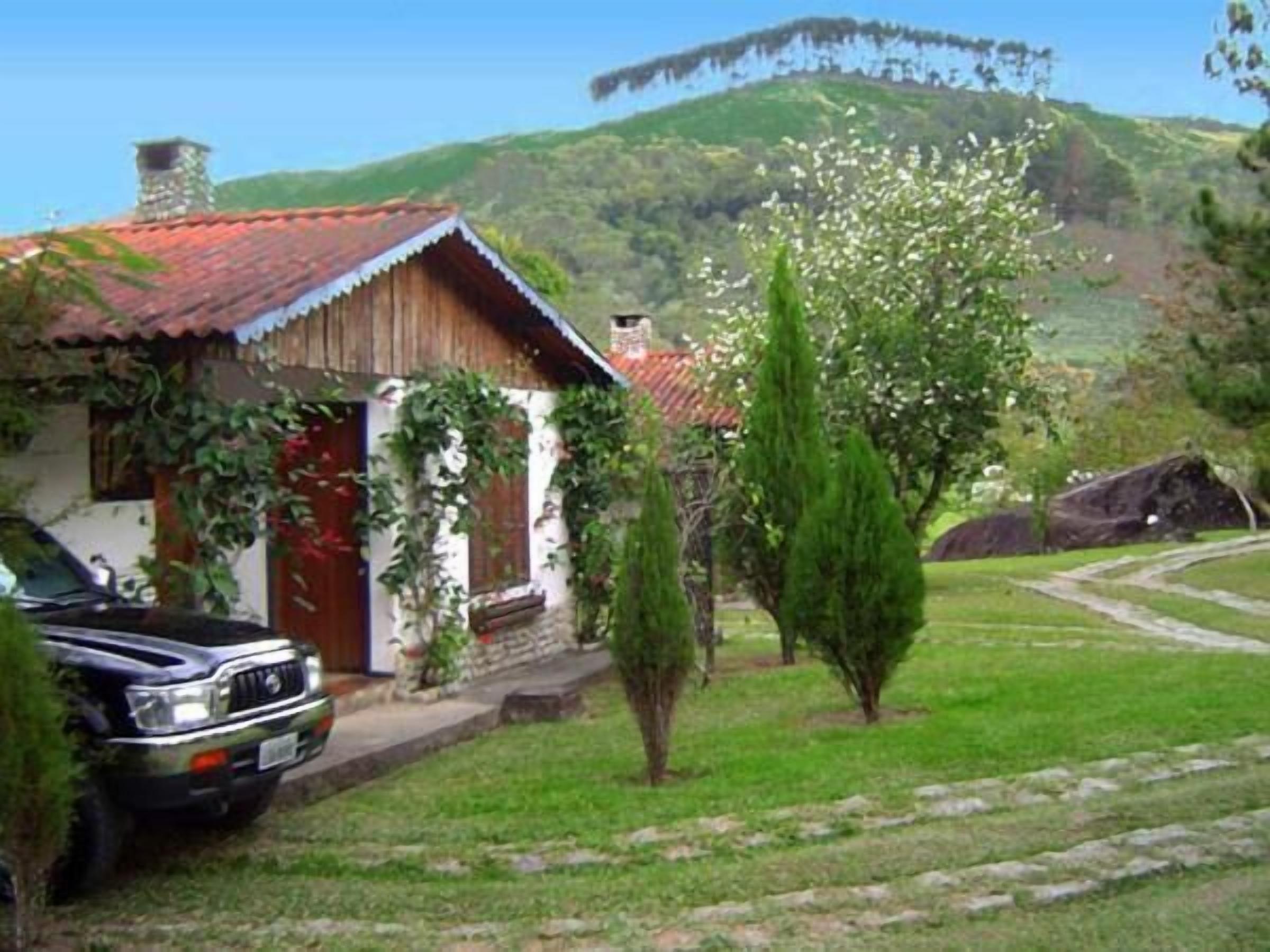 Uma casa rústica com telhado vermelho, um carro estacionado e uma paisagem verdejante com uma colina ao fundo.