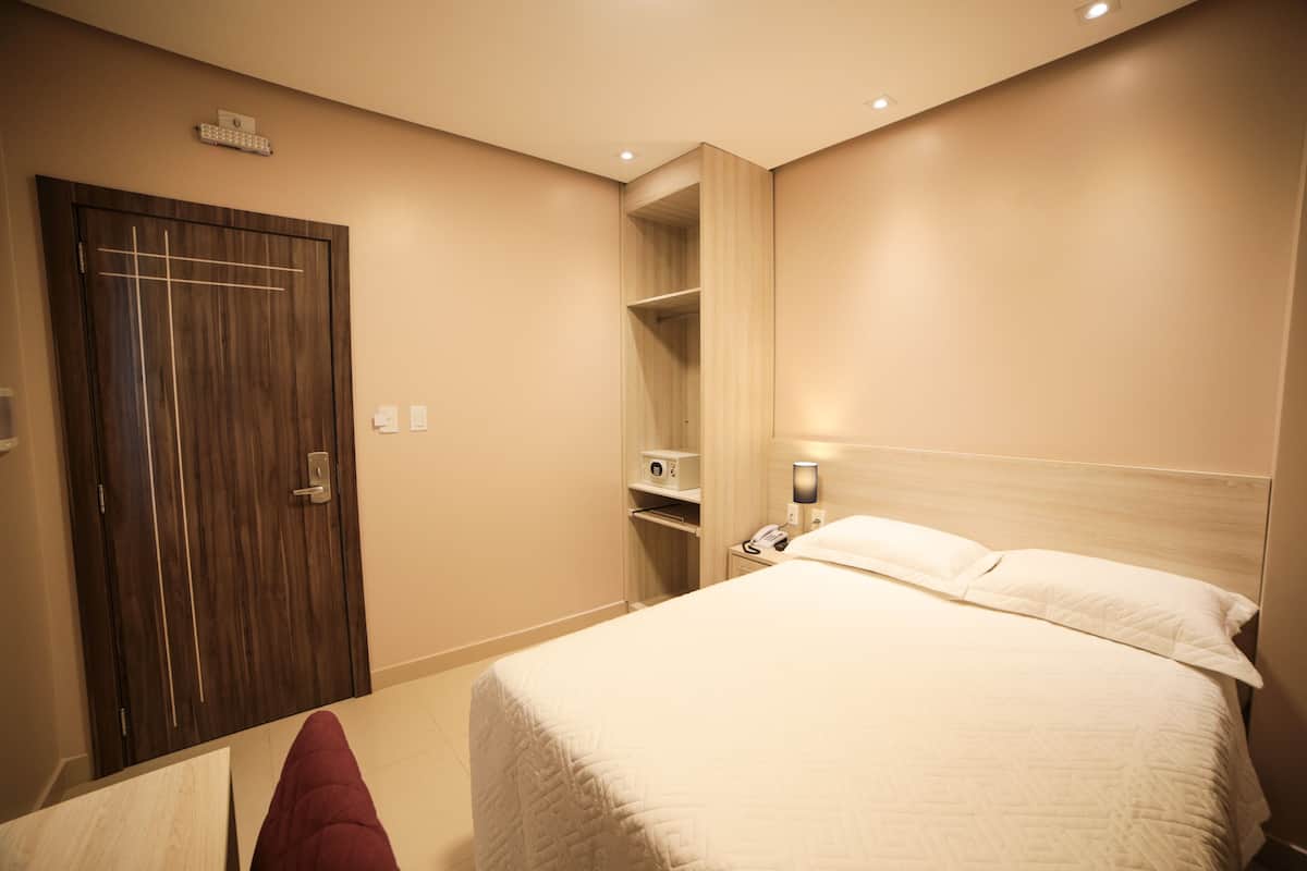 A hotel room with a wooden wardrobe, a bedside table with a lamp, and a bed with white bedding.