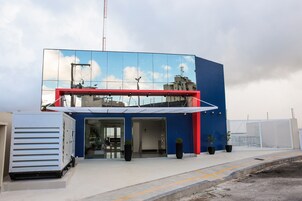 Un edificio moderno con fachada de vidrio que refleja el cielo y las nubes, un alero rojo y plantas en macetas en la entrada.