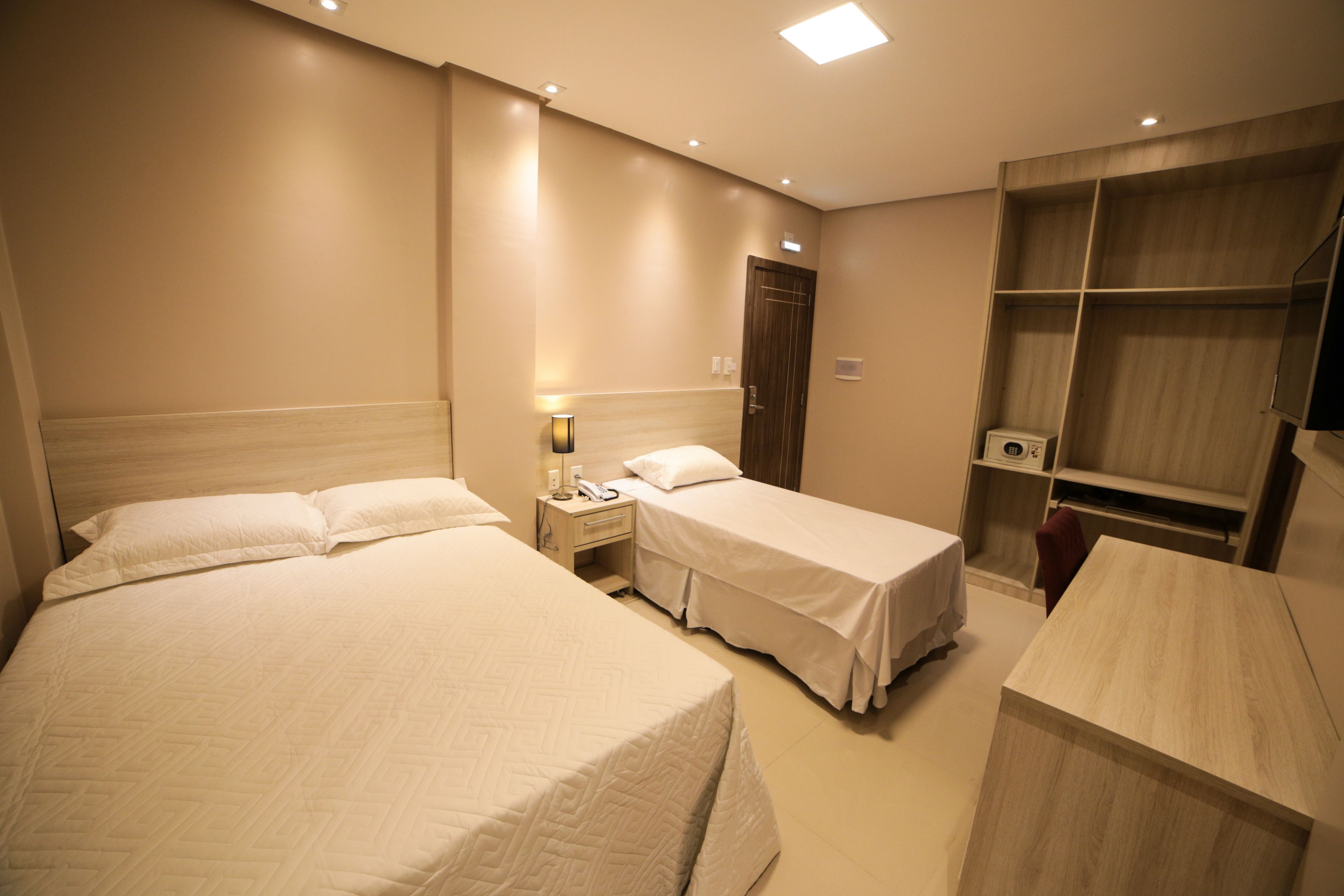 A hotel room with two beds, a bedside table with a lamp, a wardrobe, and a desk.