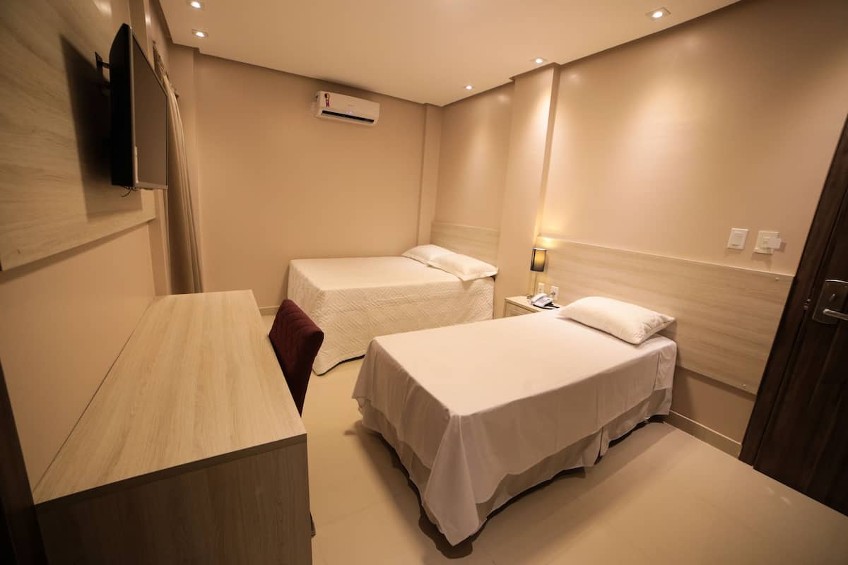 A hotel room with two single beds, a desk, a TV, an air conditioner, and a bedside lamp.