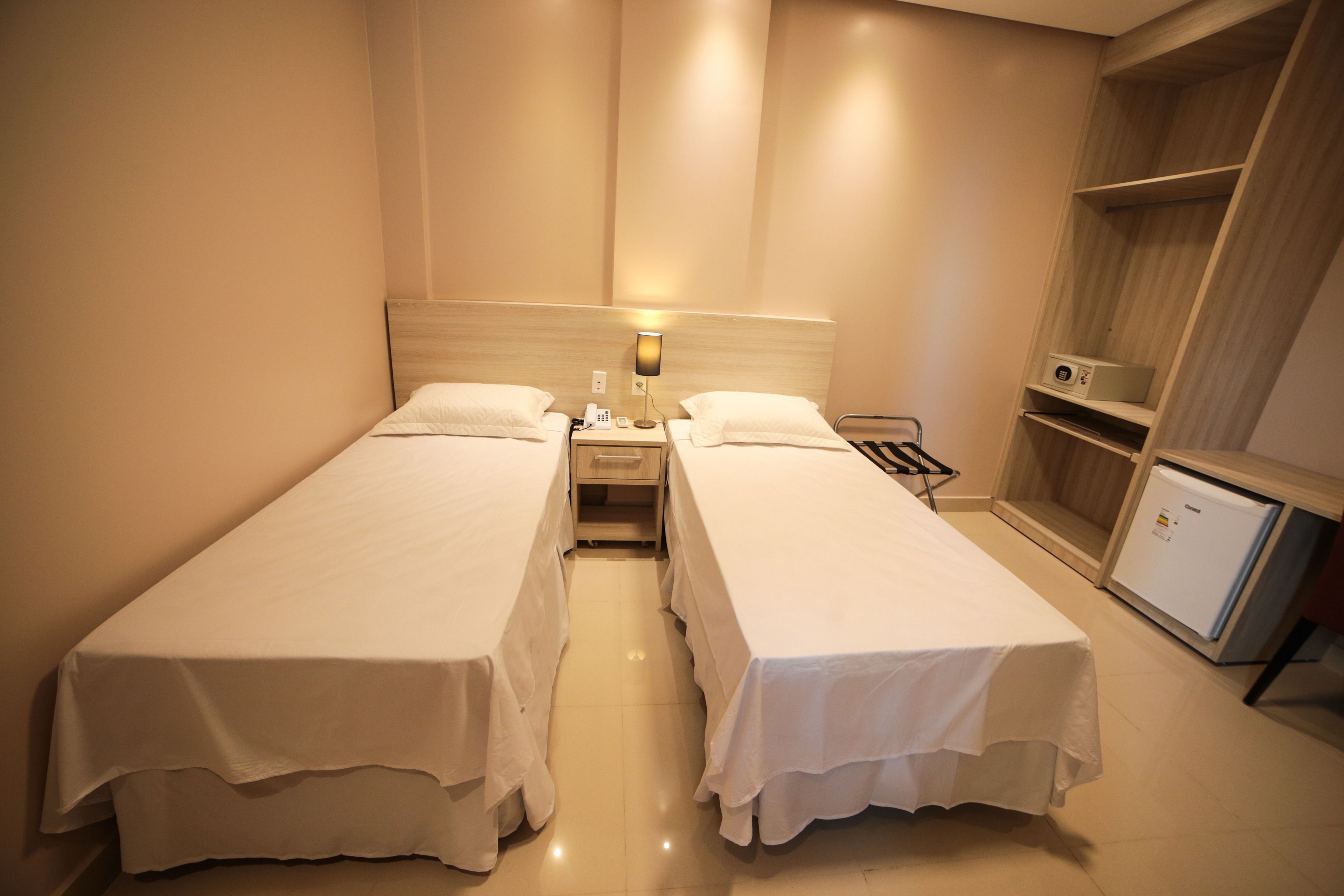 A hotel room with two single beds, a nightstand with a lamp, a mini-fridge, and a built-in wardrobe.