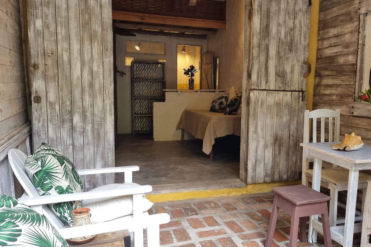 Un interior rústico con una puerta de madera, una mesita con una planta y un sillón con un cojín estampado de motivos tropicales.